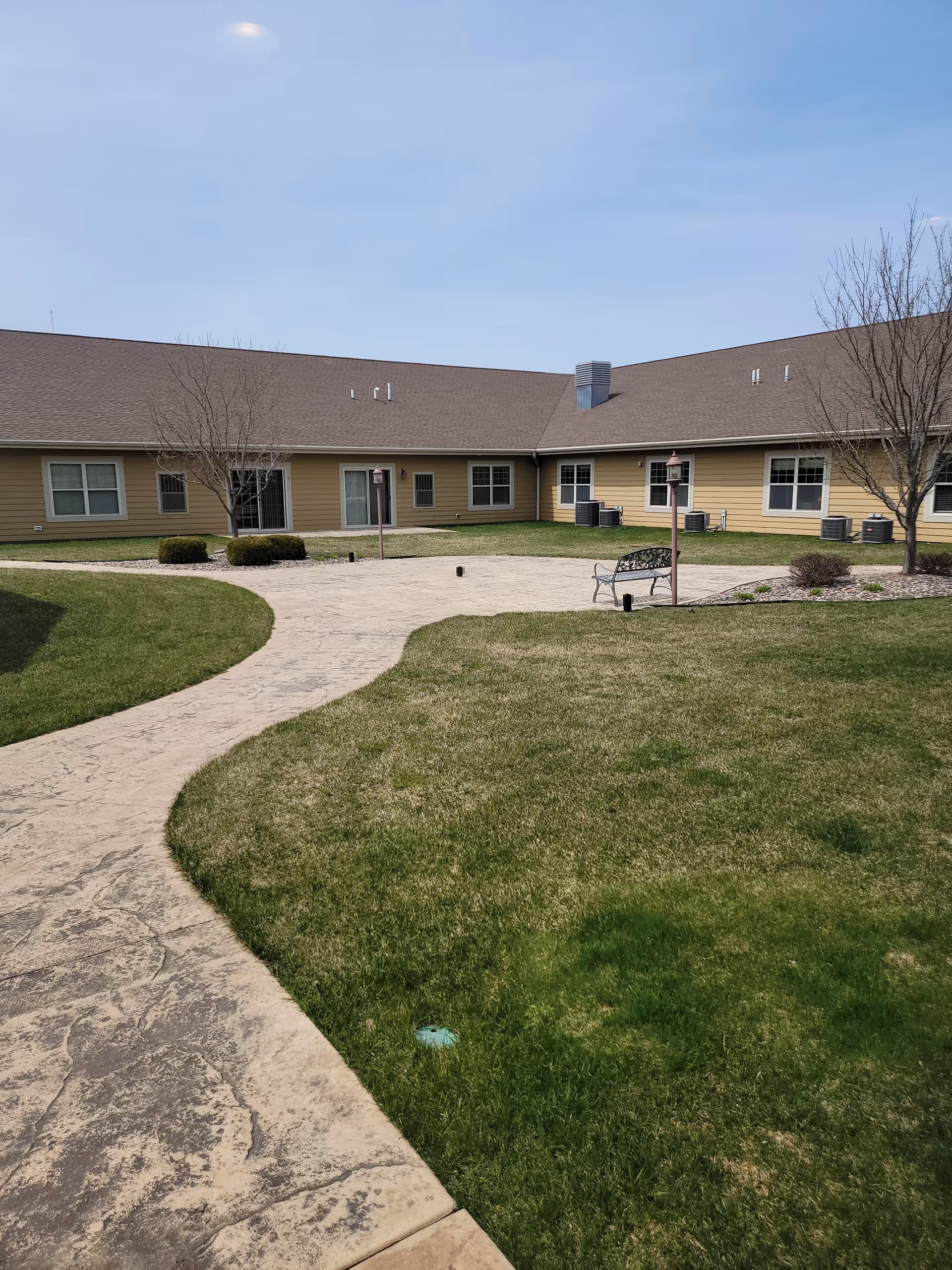Outdoor courtyard area of a senior care facility with a paved walkway, green grass, a bench, small bushes, and leafless trees. The building surrounding the courtyard has beige siding and multiple windows under a brown roof. The sky is clear and blue.
