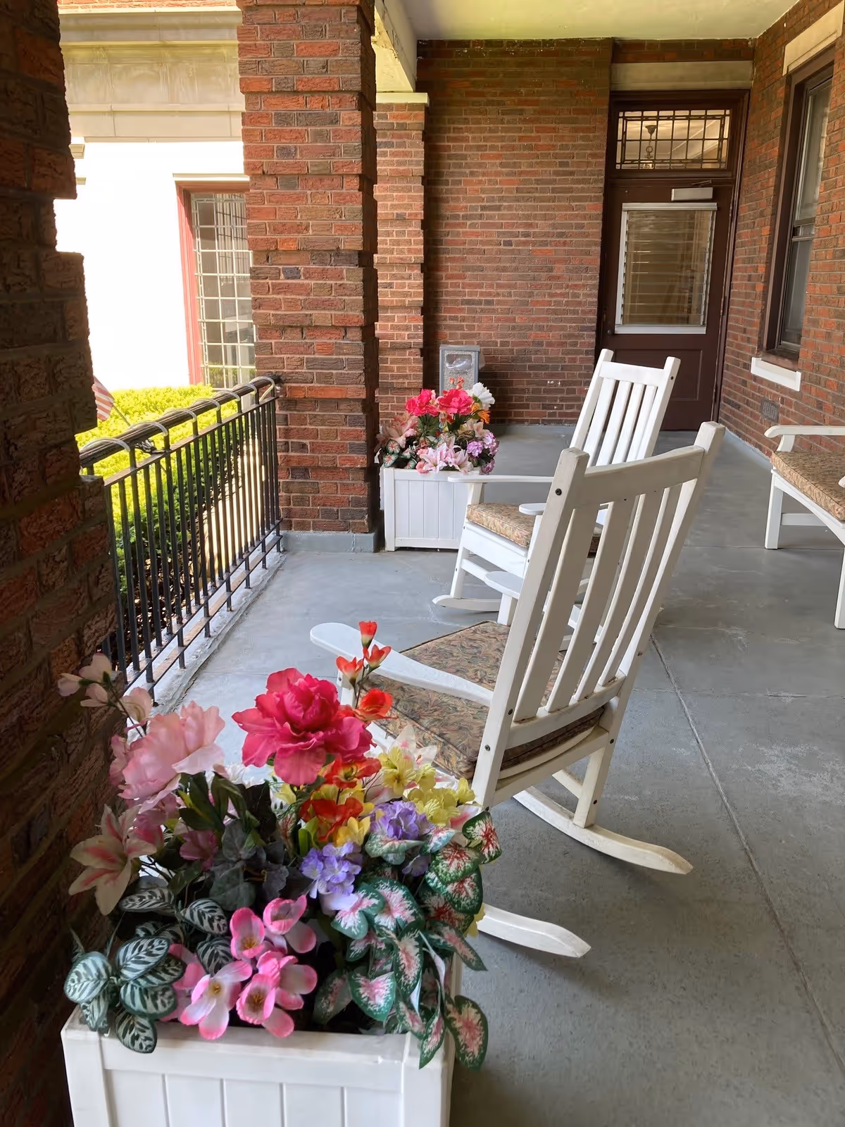 A covered outdoor porch area with two white wooden rocking chairs with cushions, surrounded by brick walls and pillars. There are white planter boxes filled with colorful artificial flowers placed near the chairs and along the porch railing. A door with a window is visible at the far end of the porch.