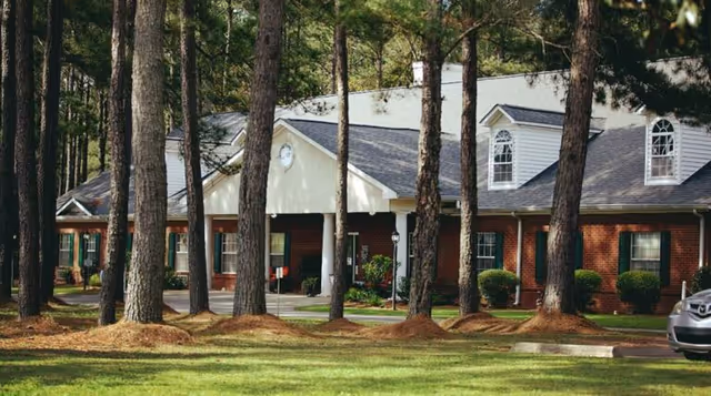 Exterior view of a senior living facility building with a brick facade, white trim, and multiple windows. The building is surrounded by tall pine trees and well-maintained grass. A car is partially visible on the right side of the image.