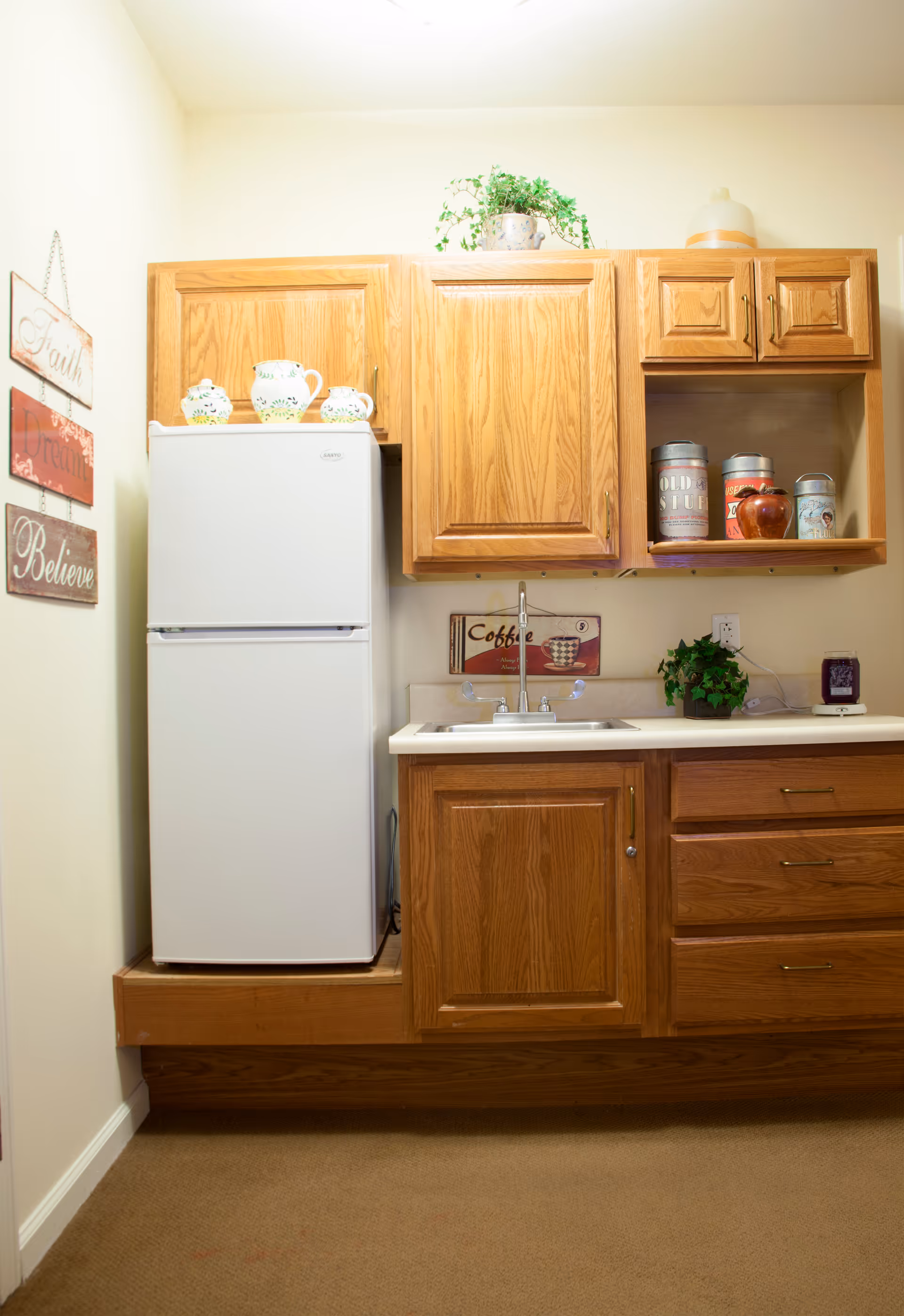 A small kitchen area with wooden cabinets, a white refrigerator, a sink, and decorative items including a potted plant, ceramic pitchers, and vintage-style canisters. On the wall to the left, there are three hanging wooden signs with the words Faith, Dream, and Believe.
