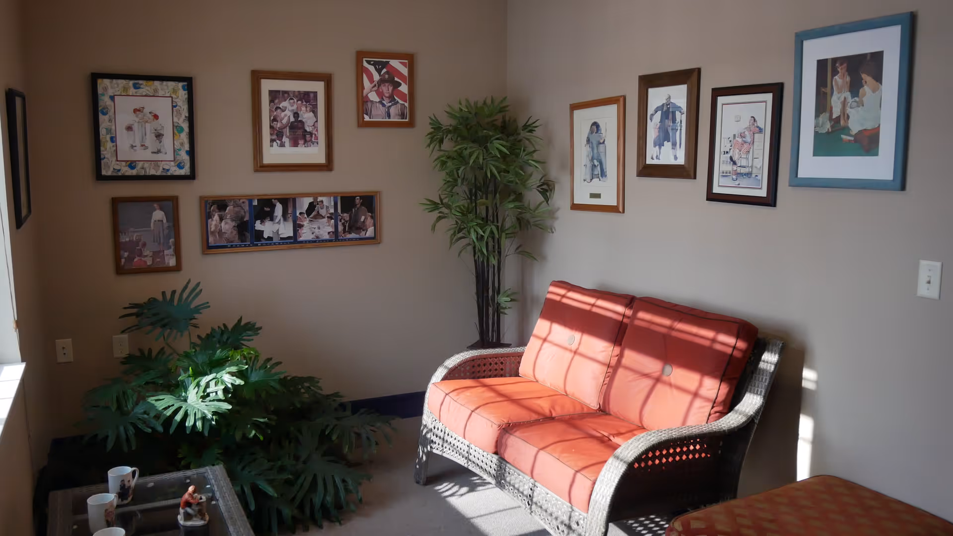 A cozy corner of a room with a wicker loveseat featuring orange cushions, a small table with two mugs and a figurine, several framed pictures on the beige walls, and two green potted plants placed near the loveseat and in the corner.