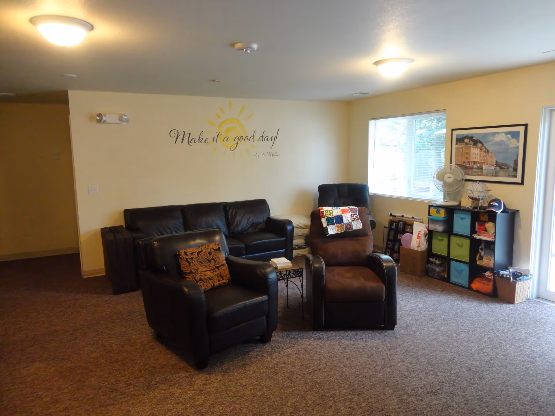 Cozy assisted-living common room with a leather sofa, two armchairs, a side table, and storage cubbies near a window.