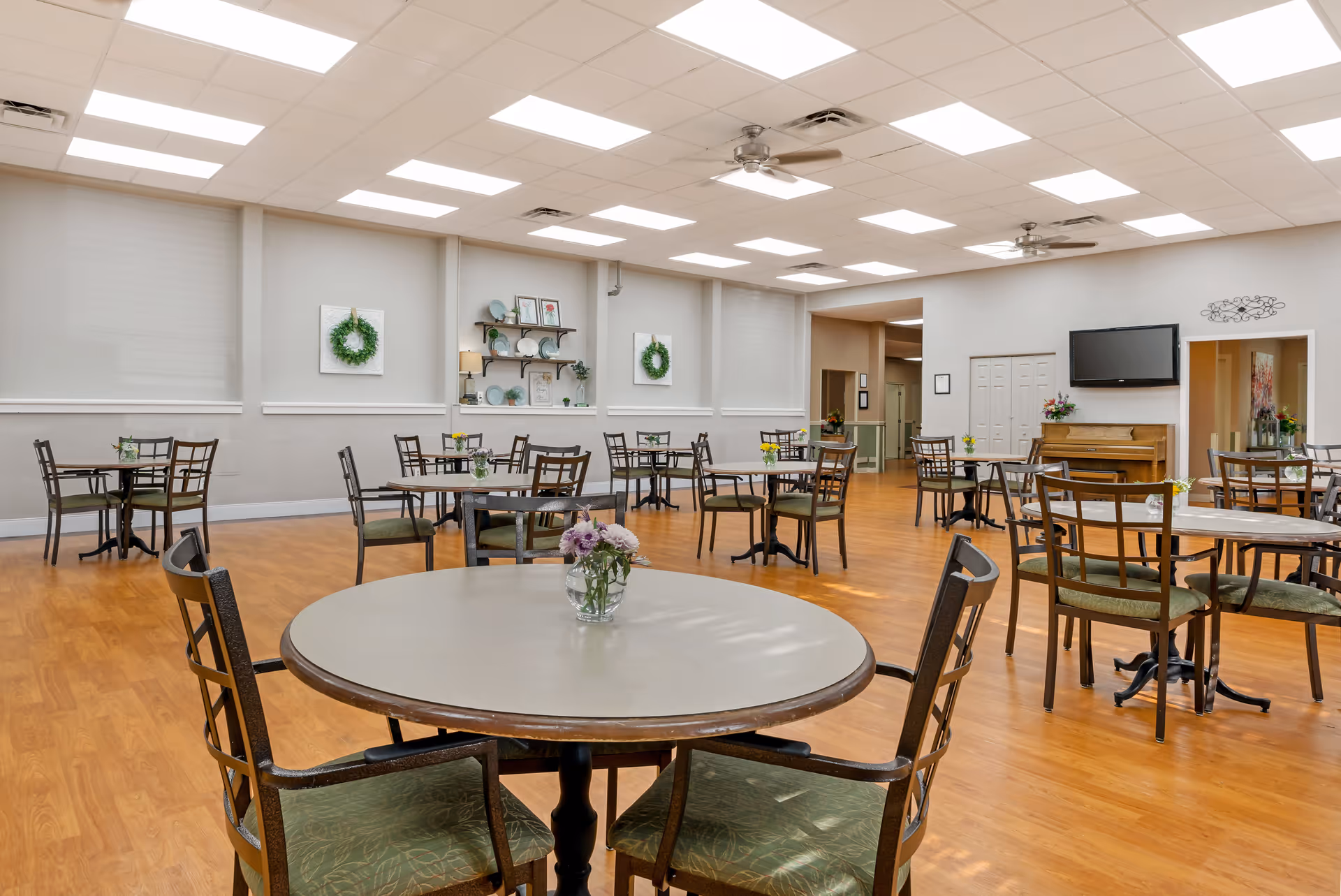 A spacious dining room with multiple round tables and chairs arranged neatly on a wooden floor. The walls are light-colored with decorative wreaths and shelves holding plates and small plants. Ceiling lights and fans illuminate the room, and a piano and wall-mounted TV are visible in the background.