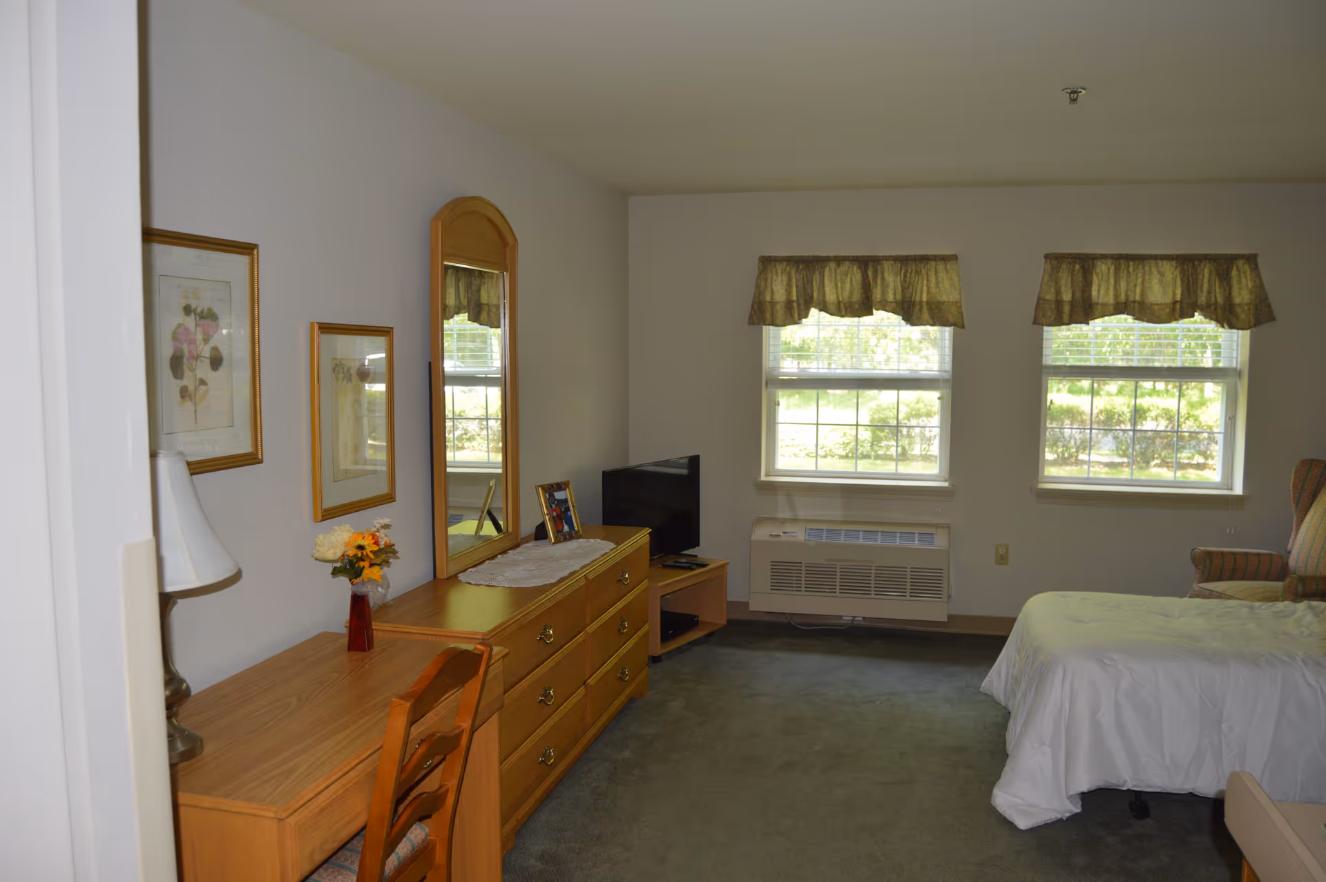 A senior living facility bedroom with a bed covered in white bedding, a wooden dresser with a mirror, a small television on a stand, two windows with valances, a wooden desk with a chair and a table lamp, and framed artwork on the wall.