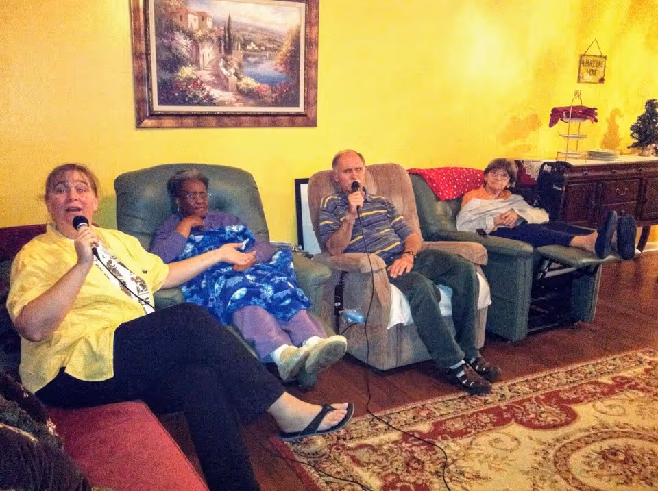 Four elderly people sitting in a living room on recliner chairs and a couch, two of them holding microphones, possibly singing karaoke. The room has yellow walls, a framed painting, a patterned rug, and a wooden cabinet with decorations.