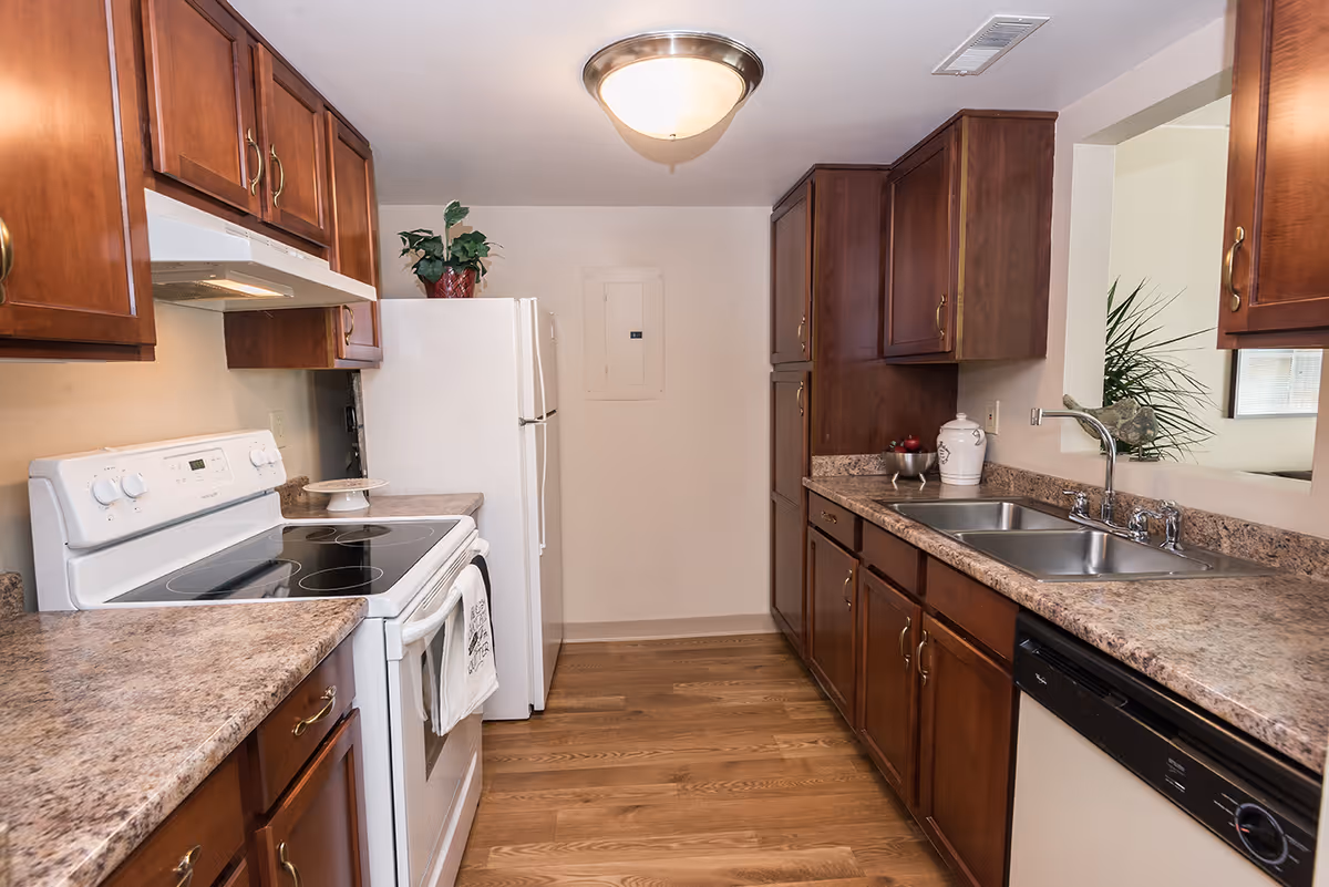 A kitchen with wooden cabinets, a white electric stove, a white refrigerator, a double sink, a dishwasher, and a countertop with decorative items including a plant and a ceramic jar. The floor is wooden and there is a ceiling light fixture.