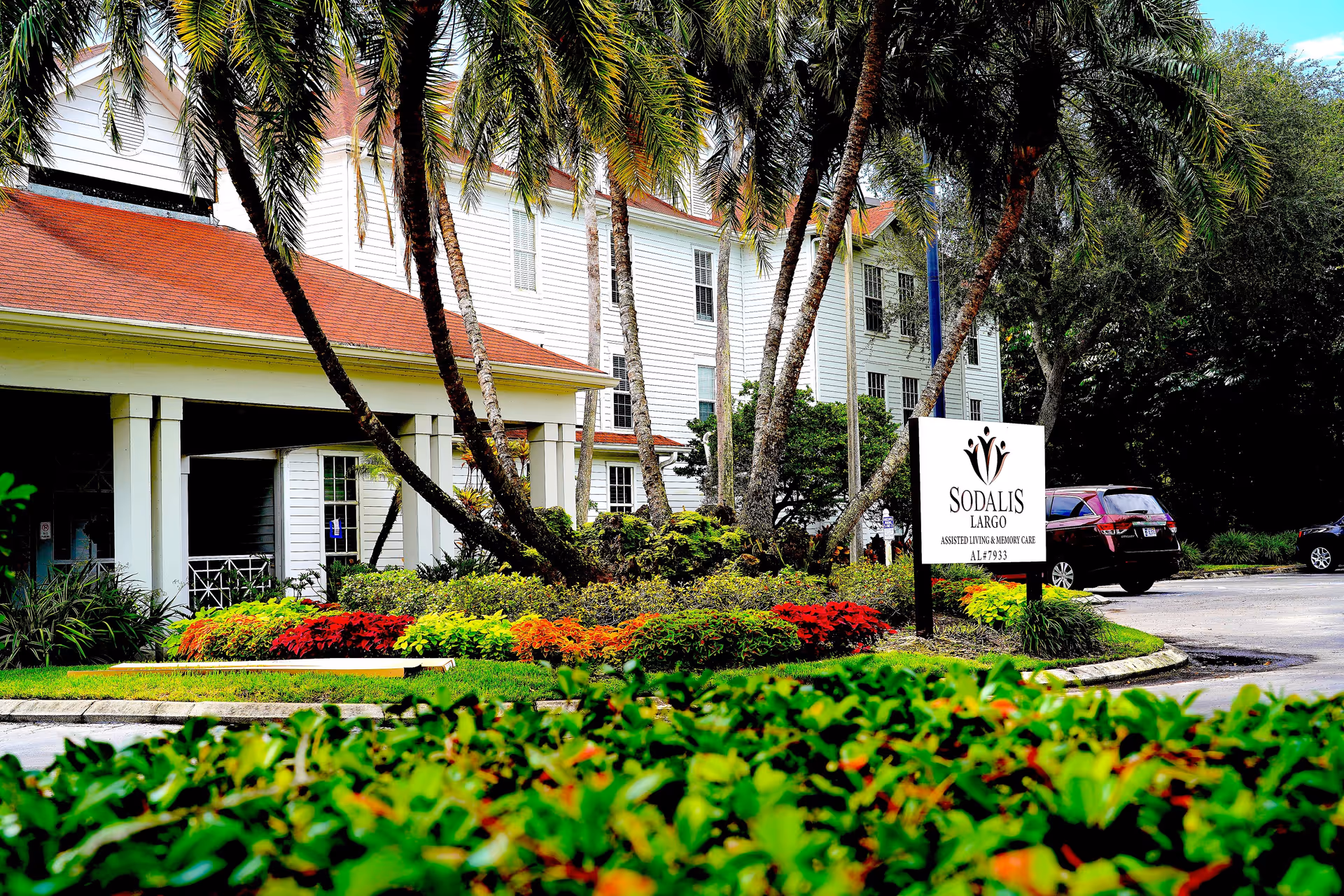 Exterior view of Sodalis of Largo facility showing a white multi-story building with a red roof, surrounded by palm trees and colorful landscaping. A sign in front reads 'Sodalis Largo Assisted Living & Memory Care AL#7933'. Several cars are parked near the entrance.