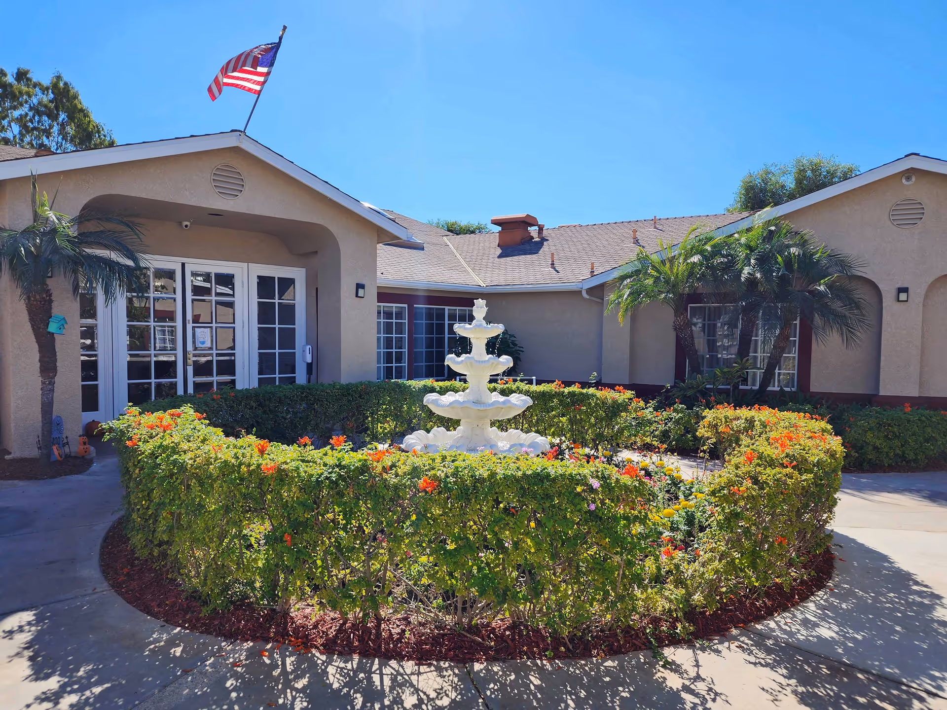 Exterior view of a single-story building with beige walls and a tiled roof, featuring a circular garden with a white tiered fountain in the center surrounded by green bushes and colorful flowers. Two palm trees flank the garden, and an American flag flies atop the building under a clear blue sky.