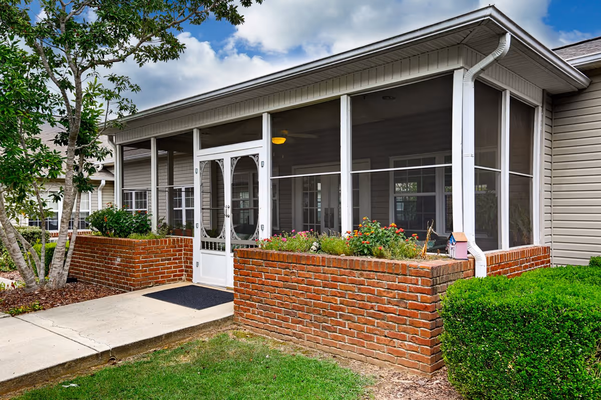 Exterior view of a screened porch attached to a building with beige siding and a brick planter box filled with flowers. There is a concrete walkway leading to a white screen door, surrounded by green bushes and a tree under a partly cloudy sky.