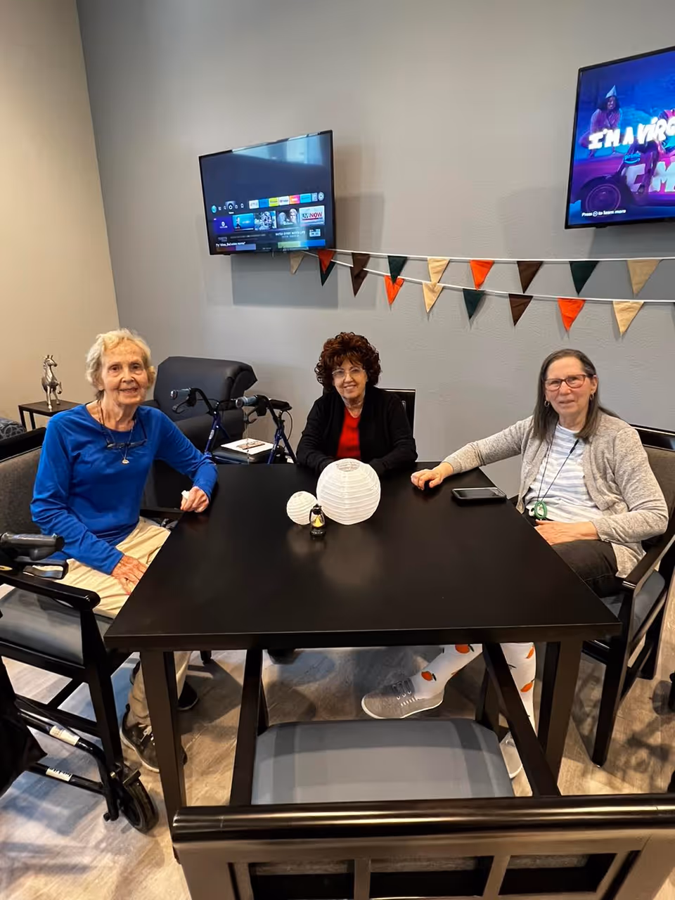Three elderly women sitting around a black table in a common room with two wall-mounted TVs and a decorative string of triangular flags on the wall behind them. One woman is wearing a blue top, another a black jacket with a red shirt, and the third a gray cardigan. There are two white paper lanterns and a small decorative item on the table.
