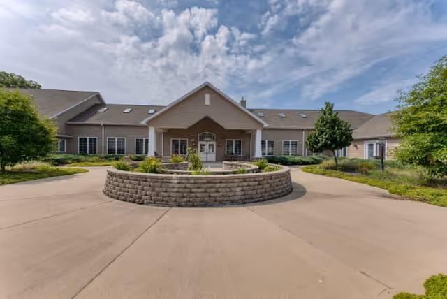 Front exterior view of a single-story building with a peaked roof and multiple windows, surrounded by landscaped greenery and a circular driveway with a stone planter in the center under a partly cloudy sky.