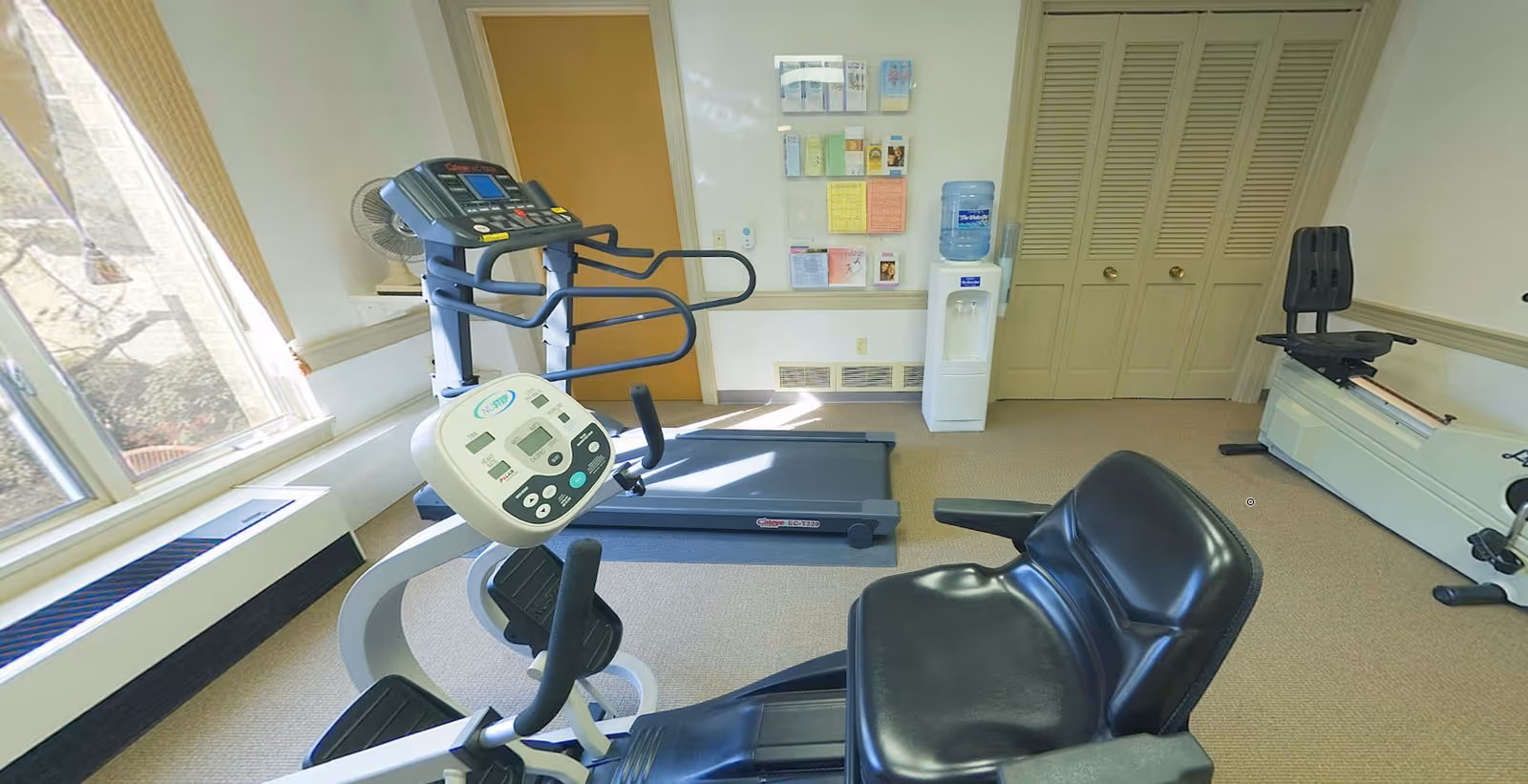 Small fitness room with exercise machines including a treadmill, recumbent bike, water cooler, and rowing machine.