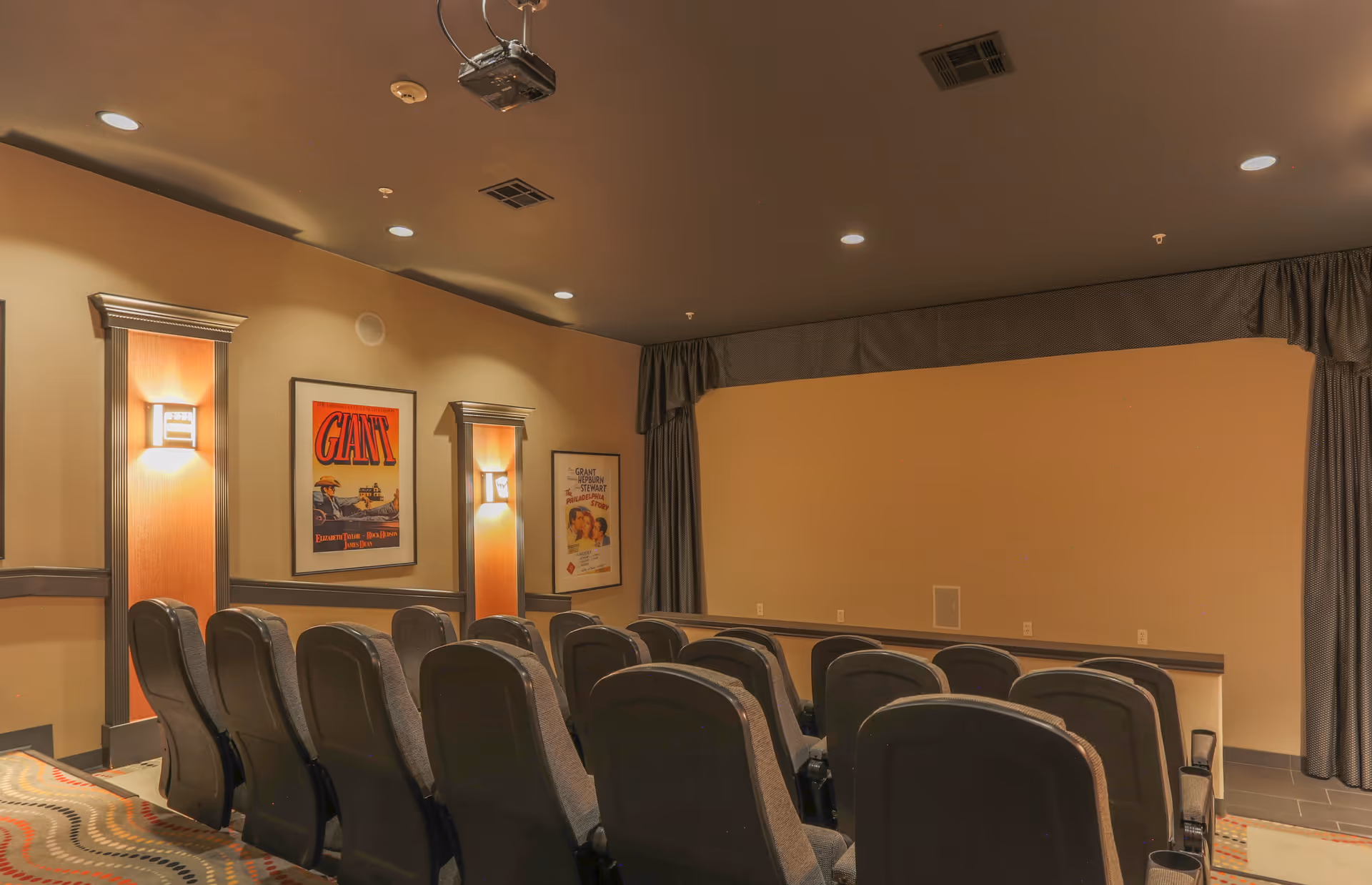 A small theater room with two rows of black cushioned seats facing a blank beige wall used as a projection screen. The room has warm lighting with wall sconces and movie posters on the walls. There is a ceiling-mounted projector and curtains framing the screen area.