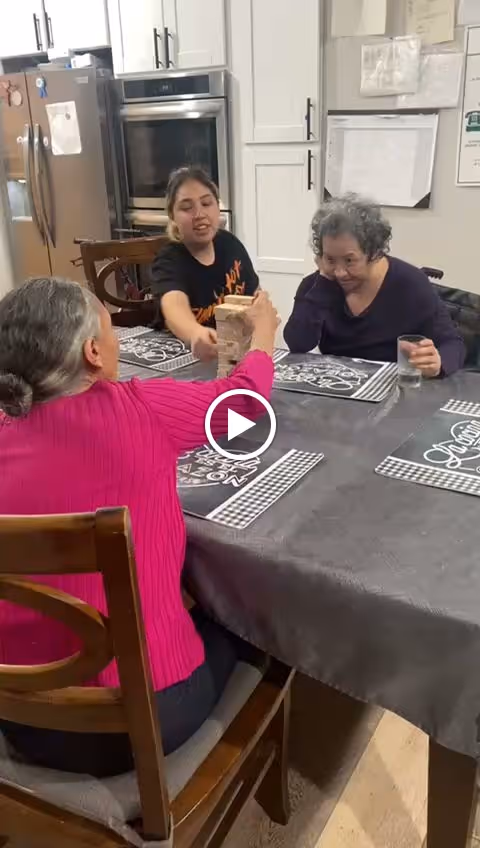 Three women sitting around a dining table playing a game of Jenga in a kitchen area. One woman is reaching to place or remove a block from the Jenga tower, while another woman watches with a glass of water in hand. The kitchen has white cabinets, a refrigerator, and an oven in the background.