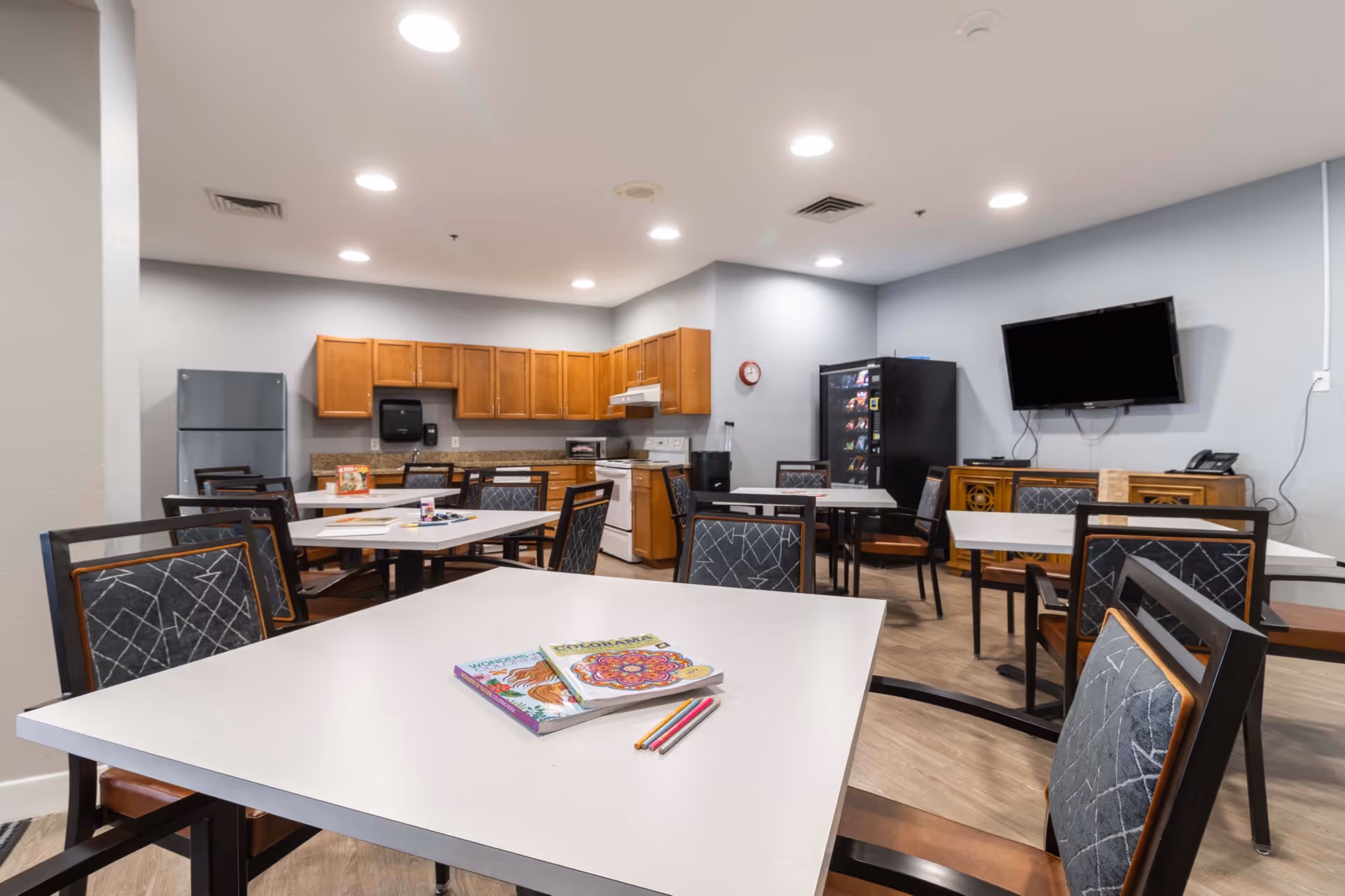 A communal room with several tables and chairs, a kitchenette with wooden cabinets, a refrigerator, stove, and microwave. There is a vending machine and a wall-mounted TV on the right side. Coloring books and colored pencils are on the table in the foreground.
