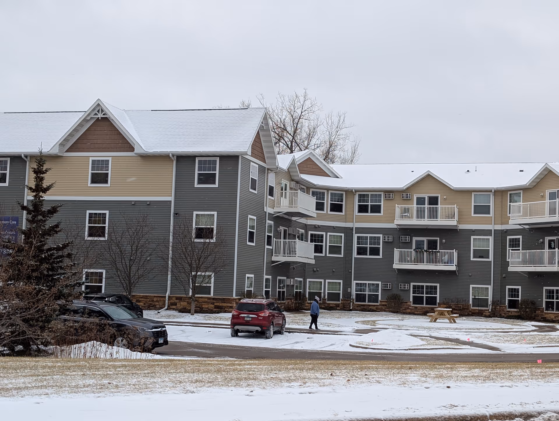 Exterior view of a multi-story senior living facility building with beige and gray siding, white balconies, and a snow-covered roof. There are a few parked cars and a person walking on a snowy path in front of the building. Leafless trees and a picnic table are visible on the snowy ground.