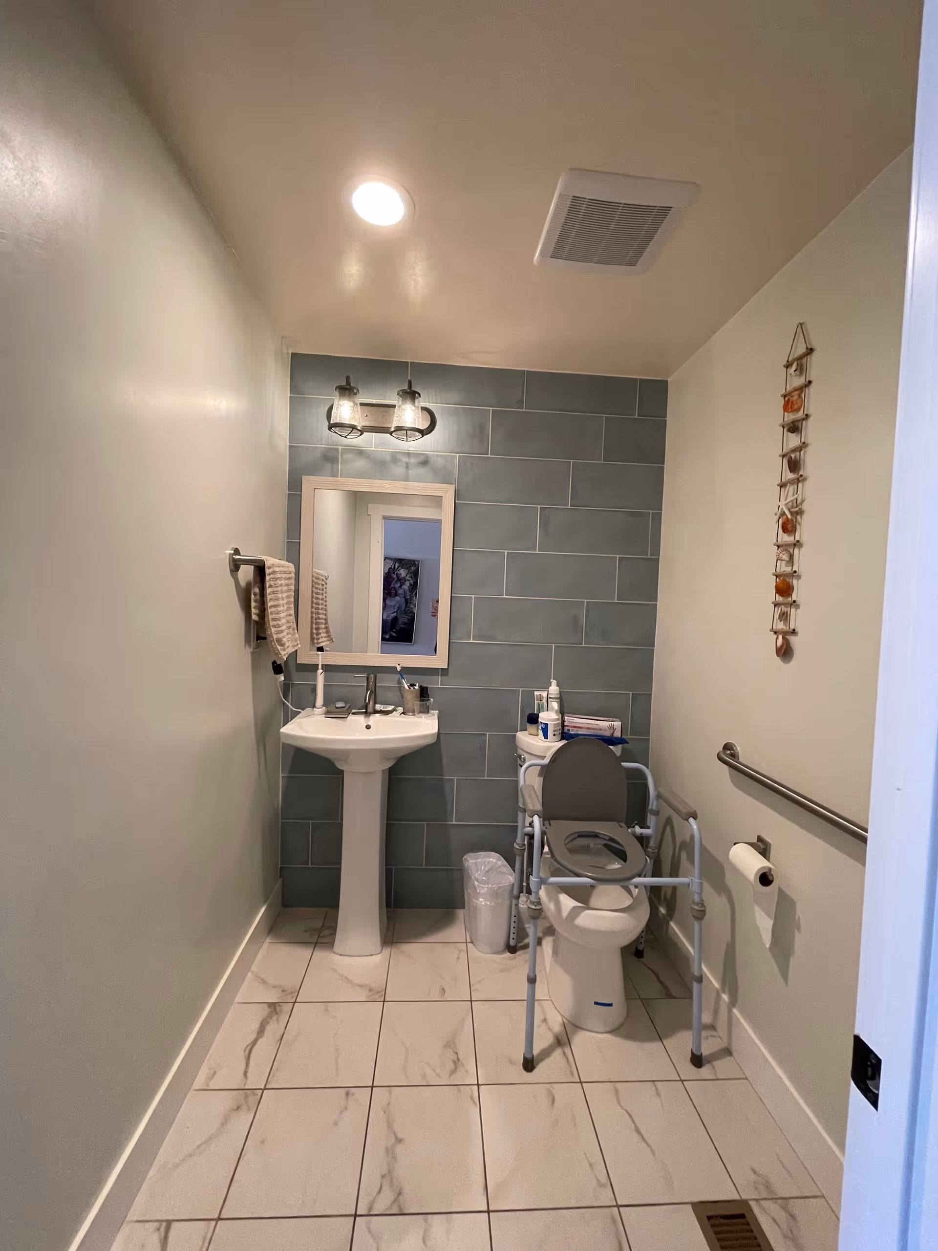 Accessible bathroom with a pedestal sink and mirror on a teal tiled accent wall, a toilet fitted with a raised support frame and grab bars, and marble-look floor tiles.