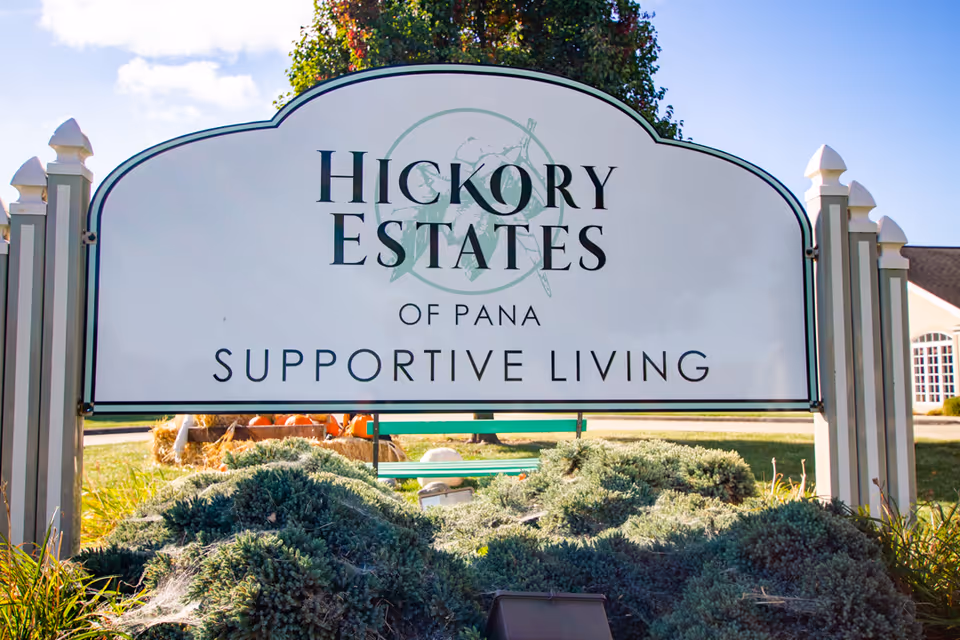Outdoor sign for Hickory Estates of Pana Supportive Living, surrounded by greenery and plants with a building partially visible in the background under a blue sky.