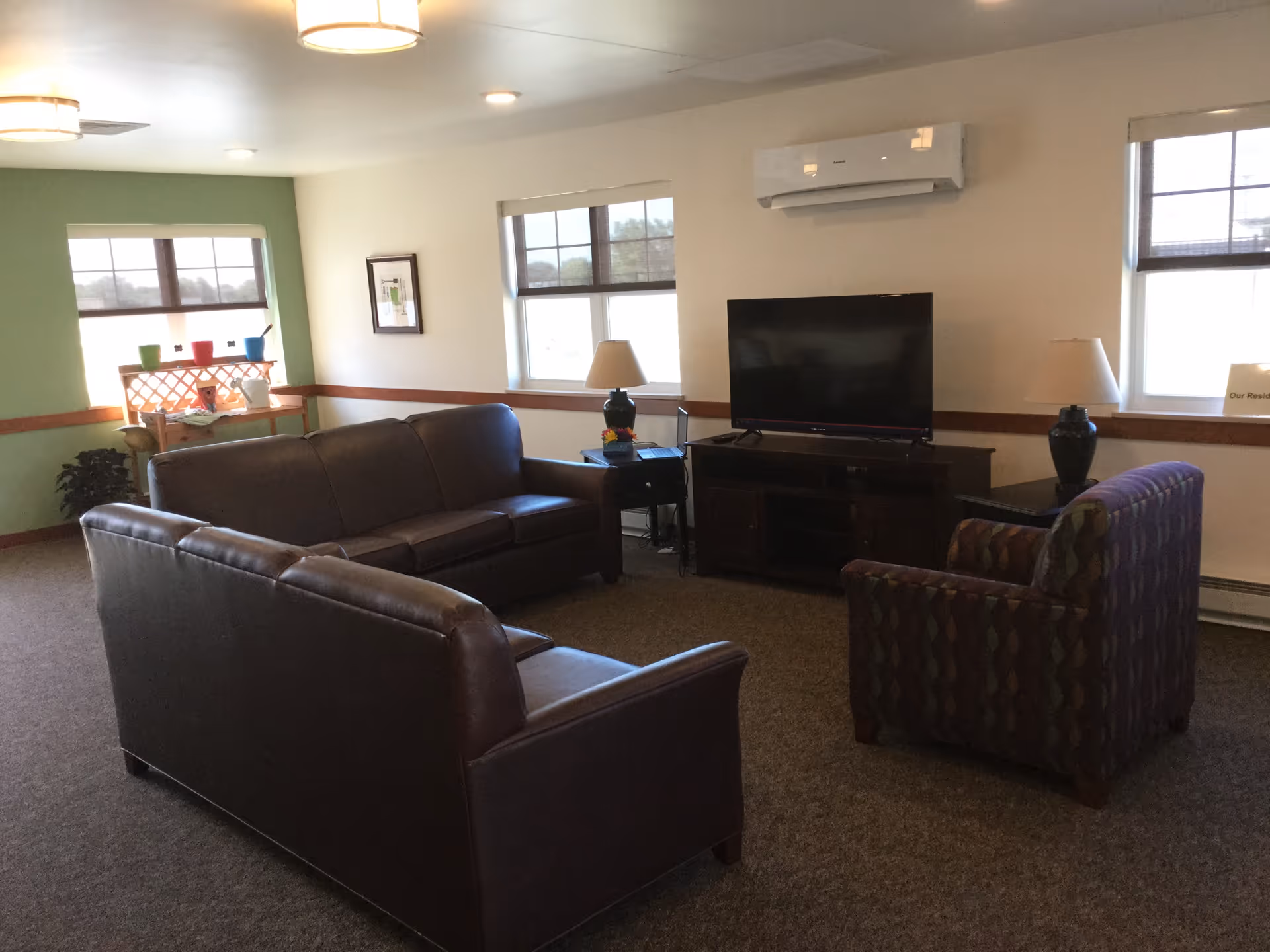 A cozy living room area with two brown leather sofas and a patterned armchair arranged around a flat-screen TV on a wooden TV stand. There are two table lamps on either side of the TV, and windows with blinds letting in natural light. A small table with colorful pots and a plant is visible in the corner.