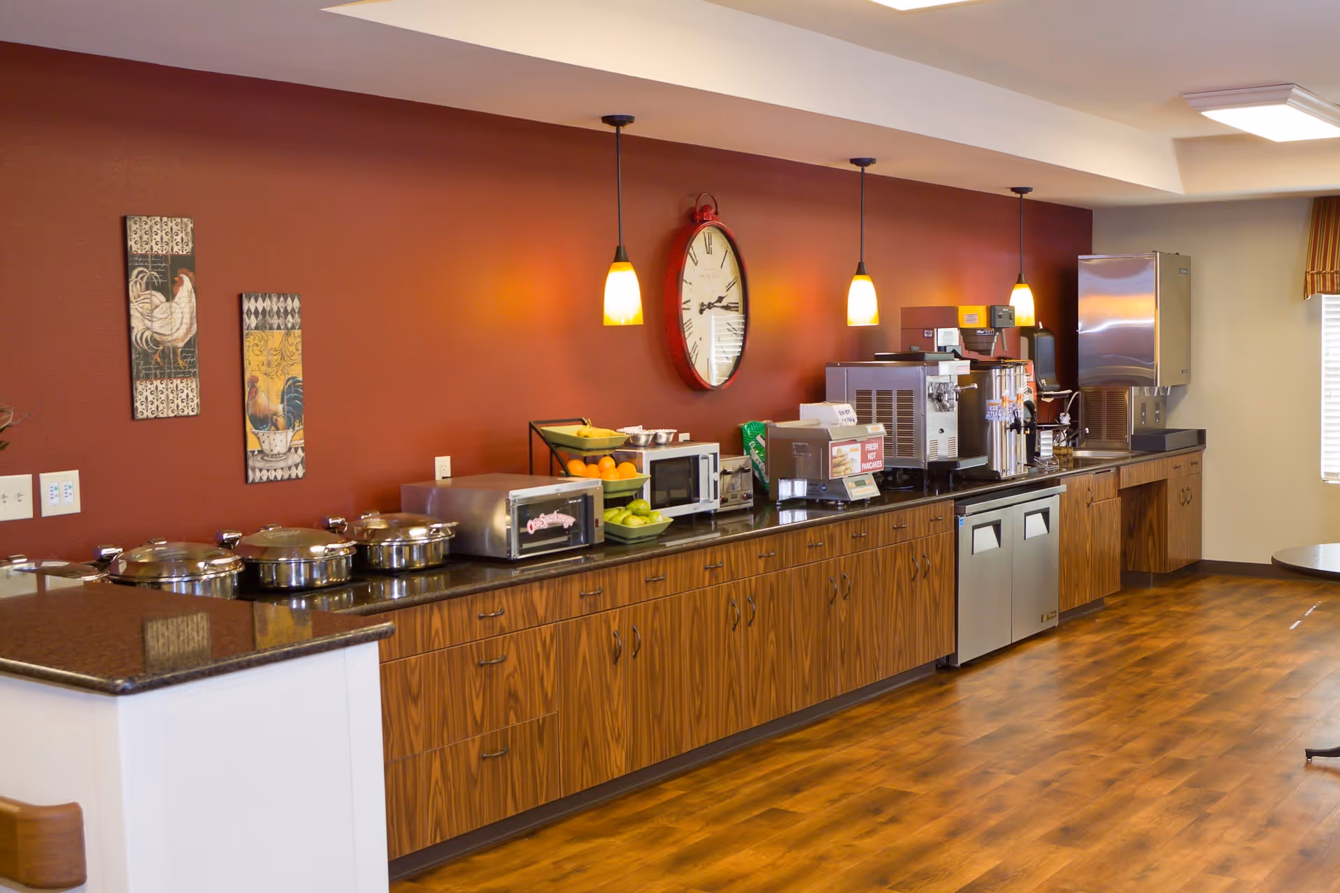 A kitchen area with wooden cabinets and a black countertop. Various kitchen appliances including a microwave, coffee machines, and a toaster oven are placed on the counter. The wall behind is painted red and decorated with two rooster-themed artworks and a large round clock. Three pendant lights hang from the ceiling, illuminating the space. The floor is wooden, and there is a window with blinds on the right side.