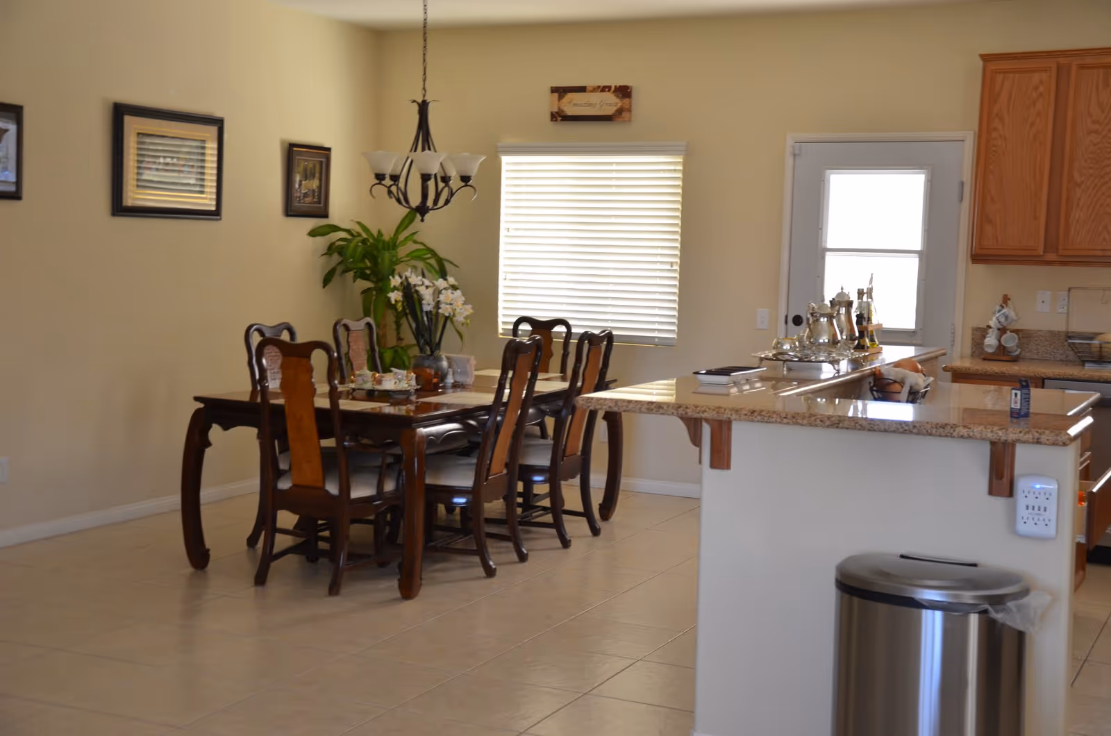A dining area with a wooden dining table and six chairs, a chandelier hanging above the table, a window with blinds, a potted plant in the corner, and a kitchen counter with various items including a silver tea set and a fruit basket. The floor is tiled and the walls are painted beige.