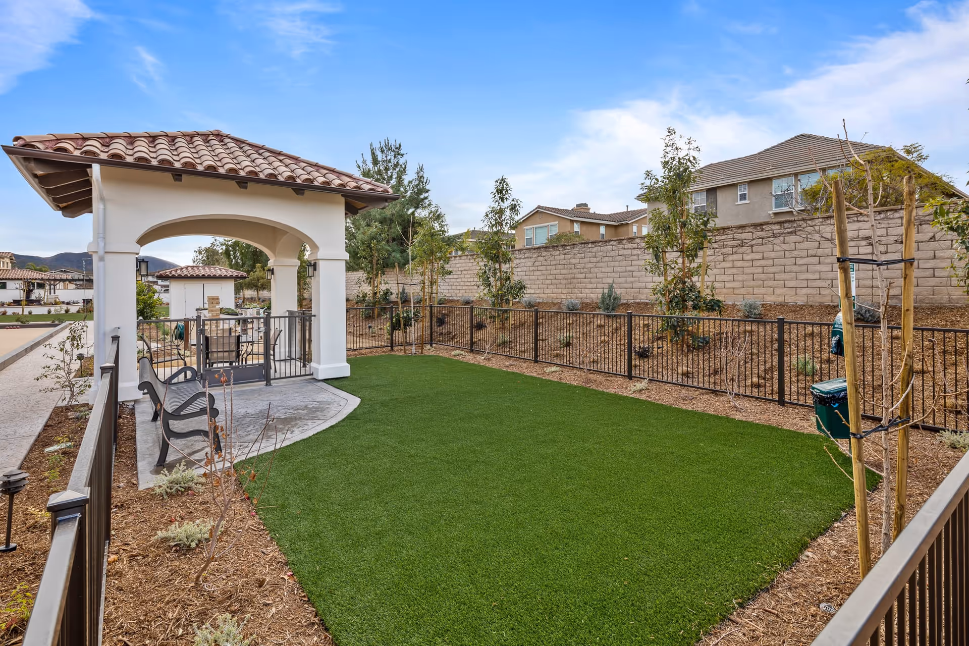 Outdoor courtyard featuring a tiled-roof gazebo, bench, fenced artificial lawn, and surrounding landscaping and nearby houses.
