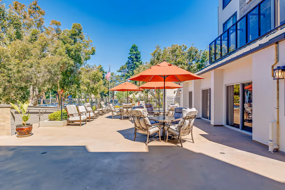 Outdoor patio area at Burlingame Senior Living with cushioned chairs and tables shaded by orange umbrellas, surrounded by trees and adjacent to a building with large windows and sliding doors under a clear blue sky.