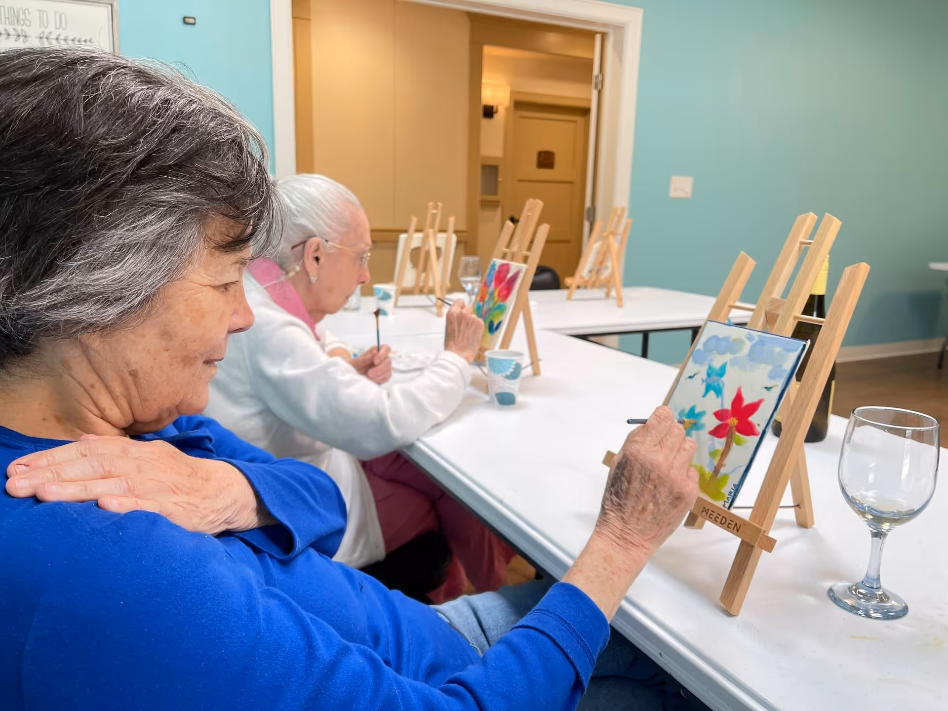 Two elderly women seated at a table in a room, painting colorful flowers on small canvases placed on wooden easels. One woman wears a blue shirt and the other a white sweater with a pink collar. The room has light blue walls and a door in the background. There are cups and a wine glass on the table.