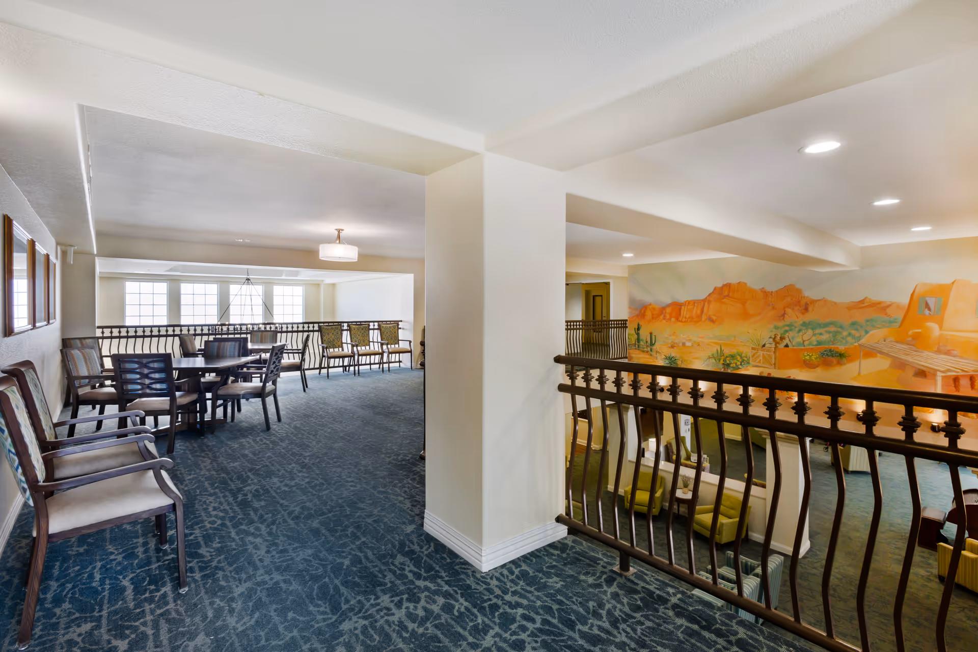 Interior view of a senior living facility common area with a seating area on the lower level and a dining or meeting area on the upper level. The upper level has several chairs and tables, carpeted floors, and a large mural of a desert landscape on the wall. The space is well-lit with ceiling lights and natural light from large windows.