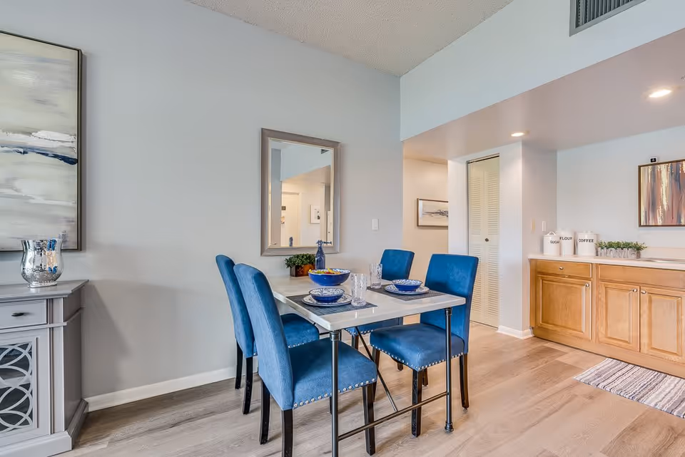 A dining area with a rectangular table set for four with blue cushioned chairs. The table has blue and white dishware, glasses, and a bowl of fruit. Behind the table is a wall with a mirror and a small plant. To the right, there is a wooden cabinet with containers labeled sugar, flour, and coffee, and a decorative 'HOME' sign. The floor is light wood, and the walls are painted light gray.