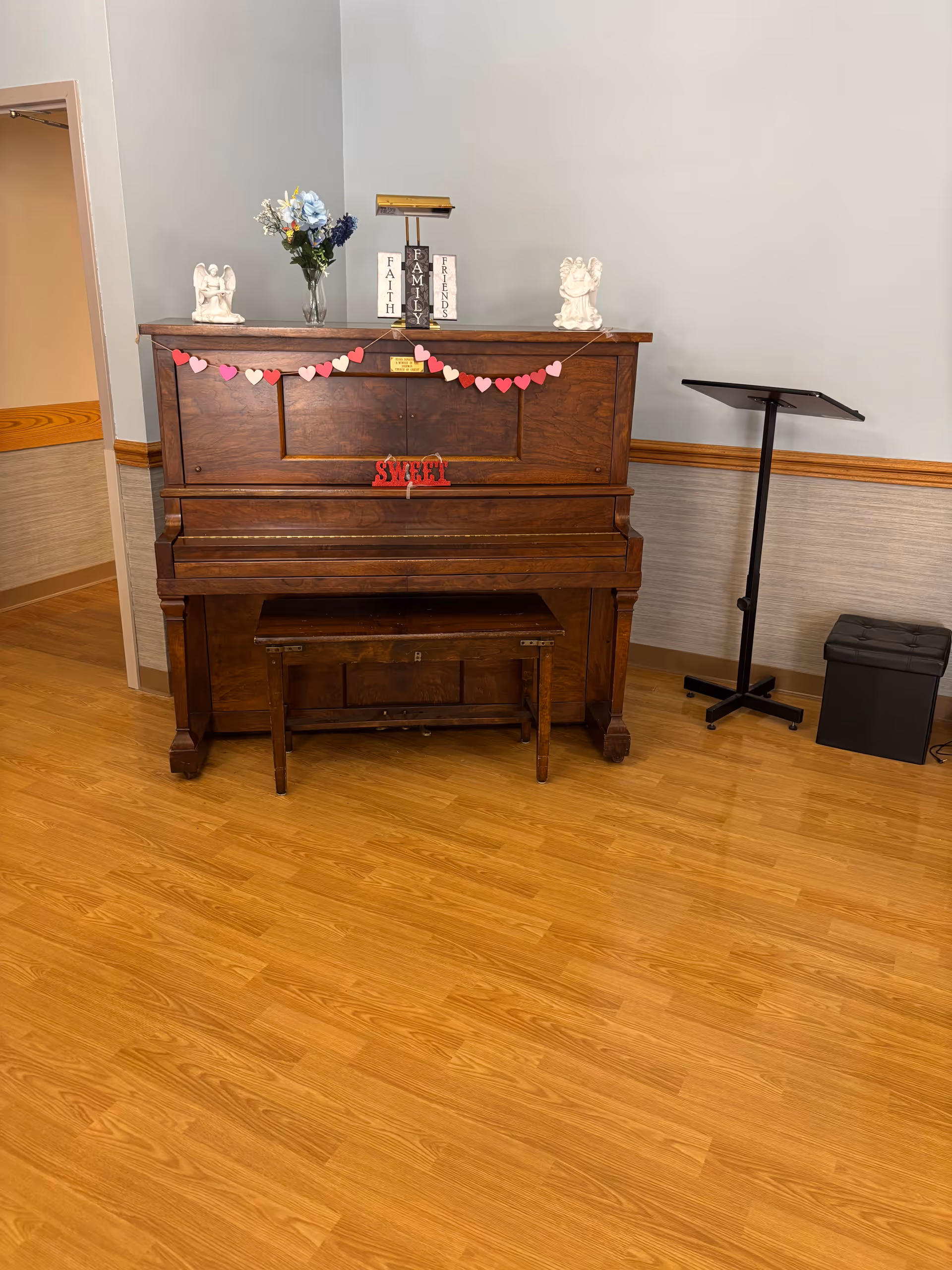 A wooden upright piano with a matching bench in front of it, decorated with a garland of red and pink paper hearts. On top of the piano are two white angel figurines, a vase with flowers, and blocks spelling out 'Faith', 'Family', and 'Friends'. To the right of the piano is a black music stand and a black storage ottoman. The room has light-colored walls with wooden trim and a wood floor.