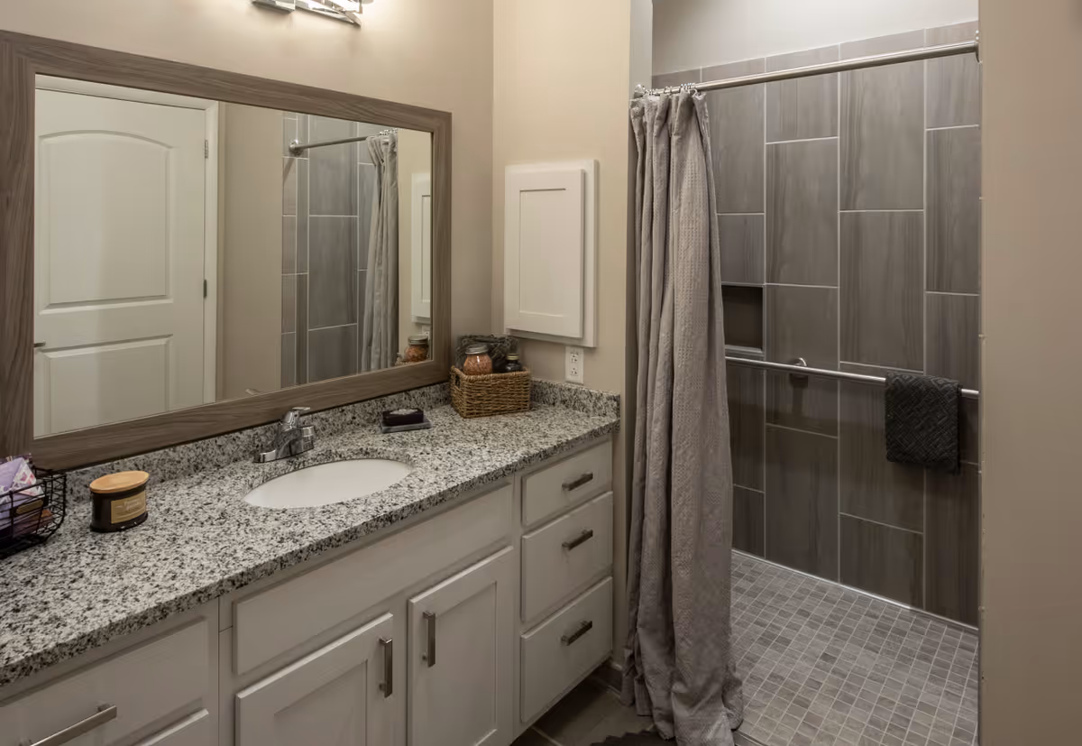 A modern bathroom featuring a granite countertop with a sink, white cabinets with silver handles, a large rectangular mirror with a wooden frame, and a walk-in shower with gray tiled walls and a gray shower curtain. There are small decorative items on the countertop including a candle and jars in a wicker basket.