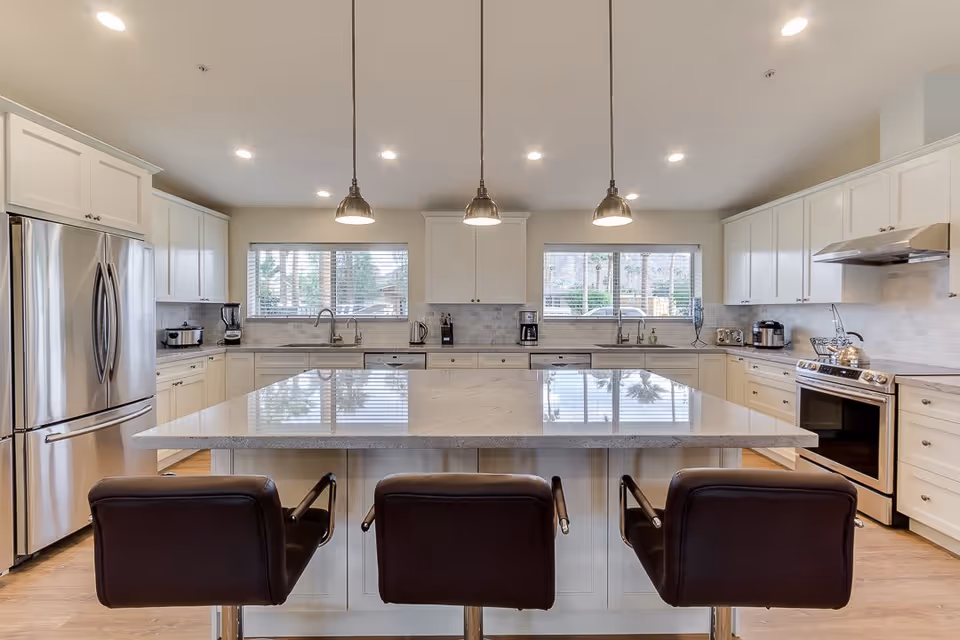 A modern kitchen with a large central island featuring a polished countertop and three black bar stools. The kitchen has white cabinetry, stainless steel appliances including a refrigerator and stove, two windows with blinds, and three pendant lights hanging above the island.