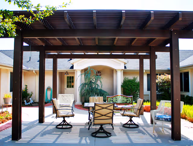 Outdoor patio area at Willow Wind Assisted Living featuring a pergola with wooden beams casting shadows on the concrete floor. Underneath, there is a table surrounded by six cushioned chairs. The patio is surrounded by a garden with plants and shrubs, and the building's exterior walls and windows are visible in the background.
