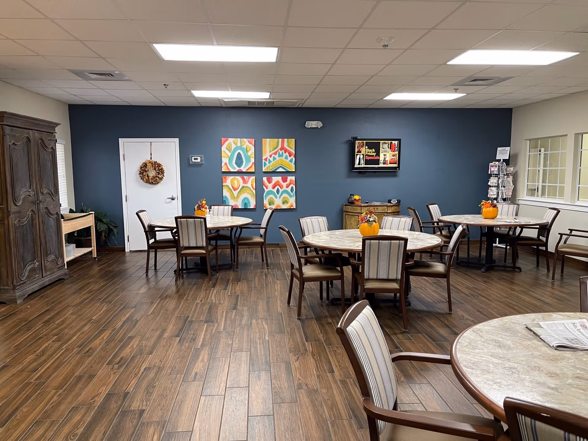 A dining room in an assisted living facility with round tables and chairs arranged neatly. Each table has a small pumpkin centerpiece with autumn-themed decorations. The room has wooden flooring, a dark blue accent wall with colorful abstract paintings, a TV mounted on the wall displaying a Black Friday Specials ad, and a cabinet and a rack with brochures near the windows.