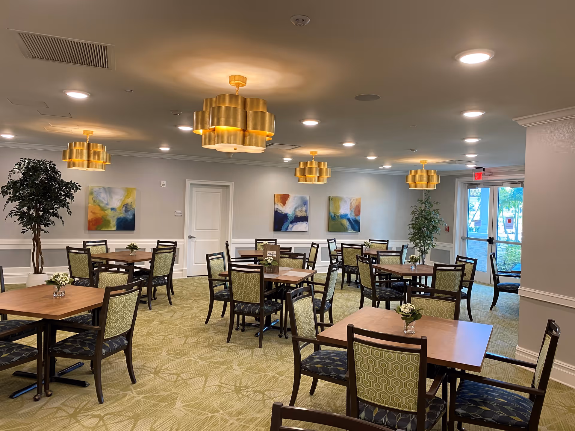 A spacious dining room in a senior living facility with multiple wooden tables and chairs arranged neatly. The room features green patterned carpet, modern gold pendant lights hanging from the ceiling, abstract paintings on the walls, and potted plants near the walls. Large glass doors provide natural light and a view of the outside.