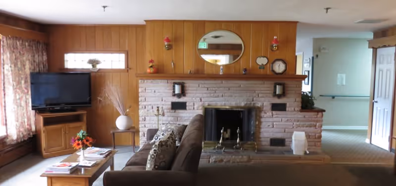 A cozy living room with wood-paneled walls, a brick fireplace with a brass screen, and a brown sofa with patterned cushions facing a wooden coffee table with magazines and a small flower arrangement. To the left, there is a TV on a wooden stand near a window with floral curtains. Above the fireplace is a round mirror and various small decorative items.
