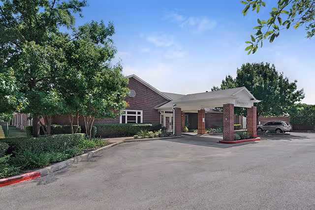 Exterior view of Brookdale North Austin facility showing a brick building with a covered entrance supported by brick columns, surrounded by trees and greenery under a clear blue sky.