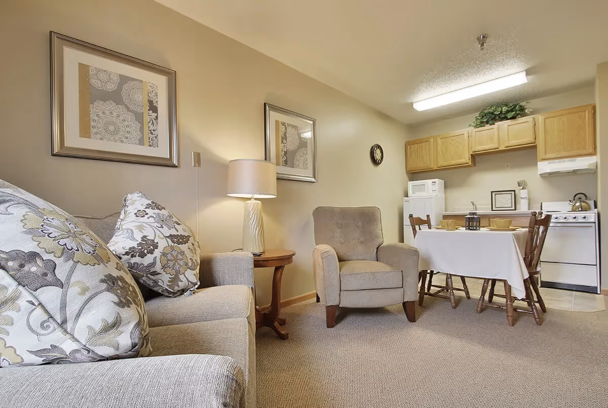 Cozy living area with a sofa and armchair, lamp and side table, small dining table, and a kitchenette with cabinets and stove.