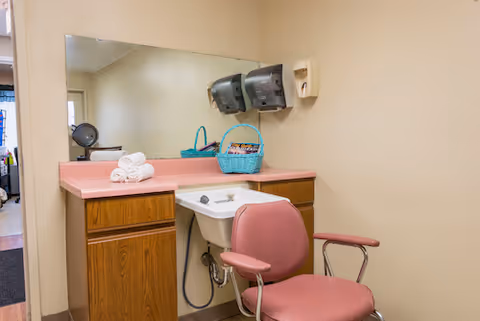 A small room with a pink cushioned chair in front of a white sink set into a wooden cabinet with a pink countertop. Above the sink is a large mirror, and mounted on the wall are two gray paper towel dispensers and a soap dispenser. A blue basket and rolled white towels are placed on the countertop.