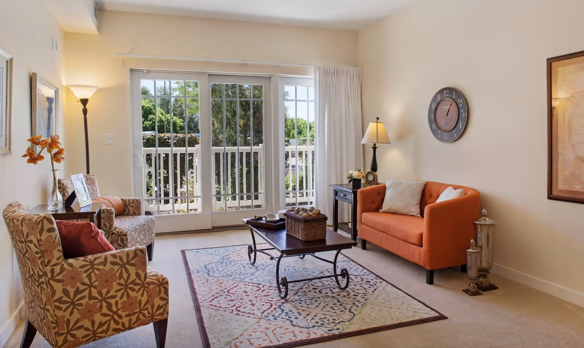 A cozy living room with a patterned armchair and an orange loveseat, a wooden coffee table on a colorful area rug, a floor lamp, a side table with a lamp and flowers, a wall clock, framed artwork, and large sliding glass doors leading to a balcony with greenery outside.