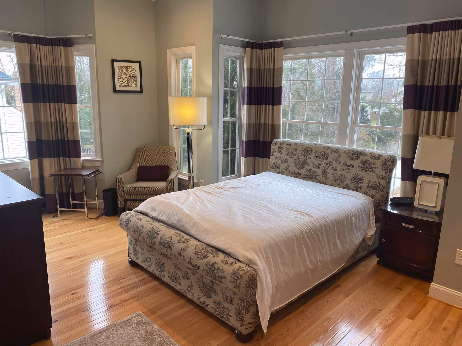 Sunlit bedroom with a patterned upholstered bed, large windows with striped curtains, an armchair and bedside tables on hardwood floors.