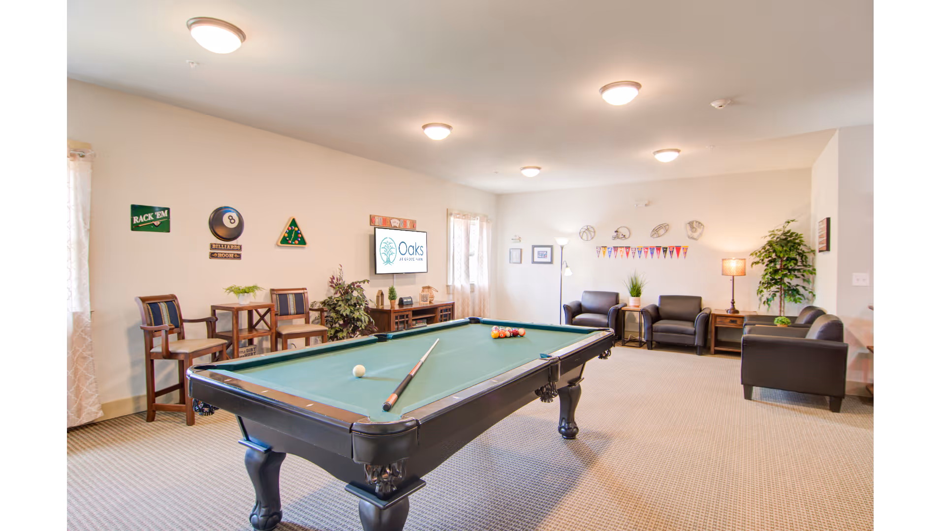 A bright and spacious game room featuring a pool table with a cue stick and balls set up for a game. The room has beige walls and carpeted floors, with several chairs and small tables along the walls. There are decorative wall hangings related to billiards and sports, a flat-screen TV mounted on the wall displaying the Oaks at Grove Park logo, and comfortable seating including armchairs and a sofa. The room is well-lit with ceiling lights and a floor lamp, and there are plants adding a touch of greenery.