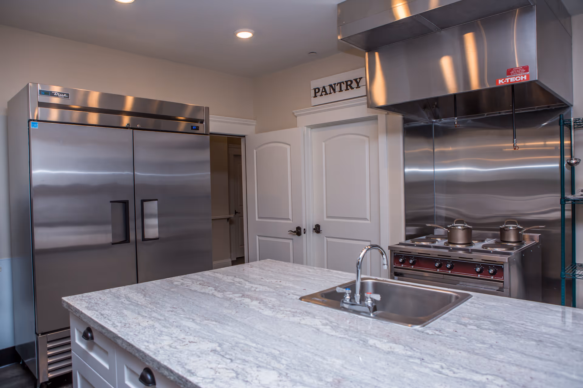 Modern kitchen area with a large stainless steel refrigerator, a stove with two pots on it, a stainless steel range hood, a marble countertop island with a sink and faucet, and two white doors labeled 'PANTRY'.