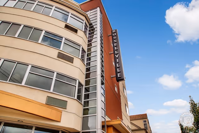 Exterior view of a multi-story building with curved beige and red walls, large windows, and a vertical sign reading 'RUSSELLVILLE' against a blue sky with scattered clouds.