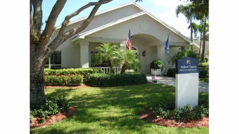 Exterior view of Arden Courts - ProMedica Memory Care Community in Delray Beach, showing a single-story building with a well-maintained lawn, shrubs, palm trees, and two flags near the entrance.