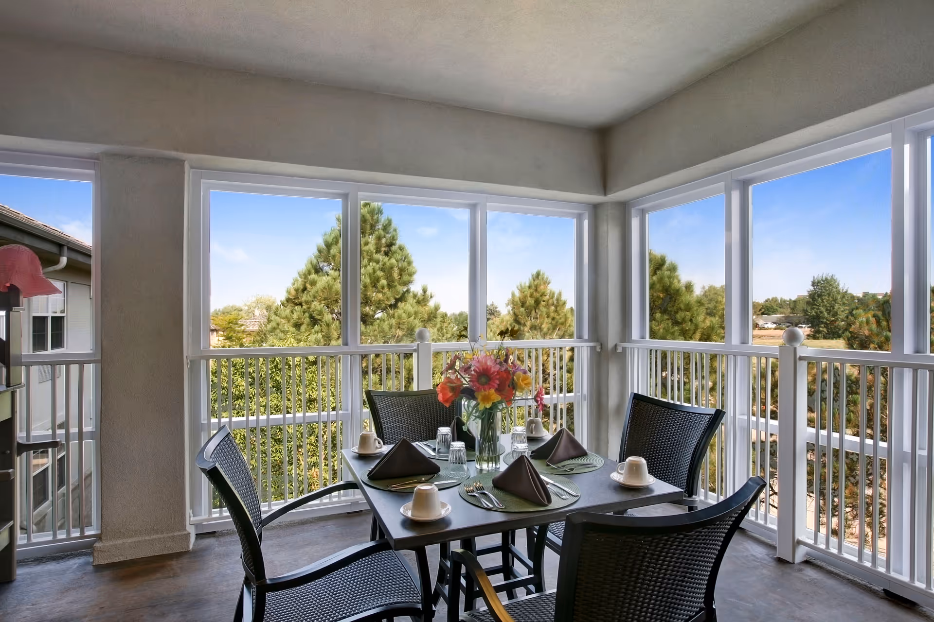 Sunlit enclosed balcony dining area with a table set for four and views of trees through large windows.