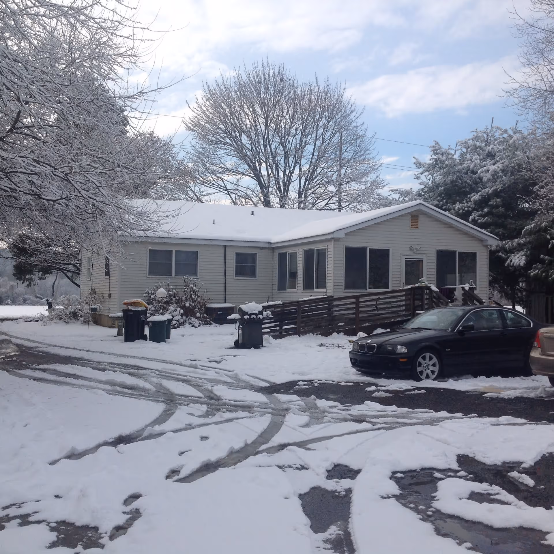 Single-story house covered with snow, surrounded by snow-covered trees and a snowy driveway with tire tracks. There are trash bins near the house and two parked cars in the driveway.