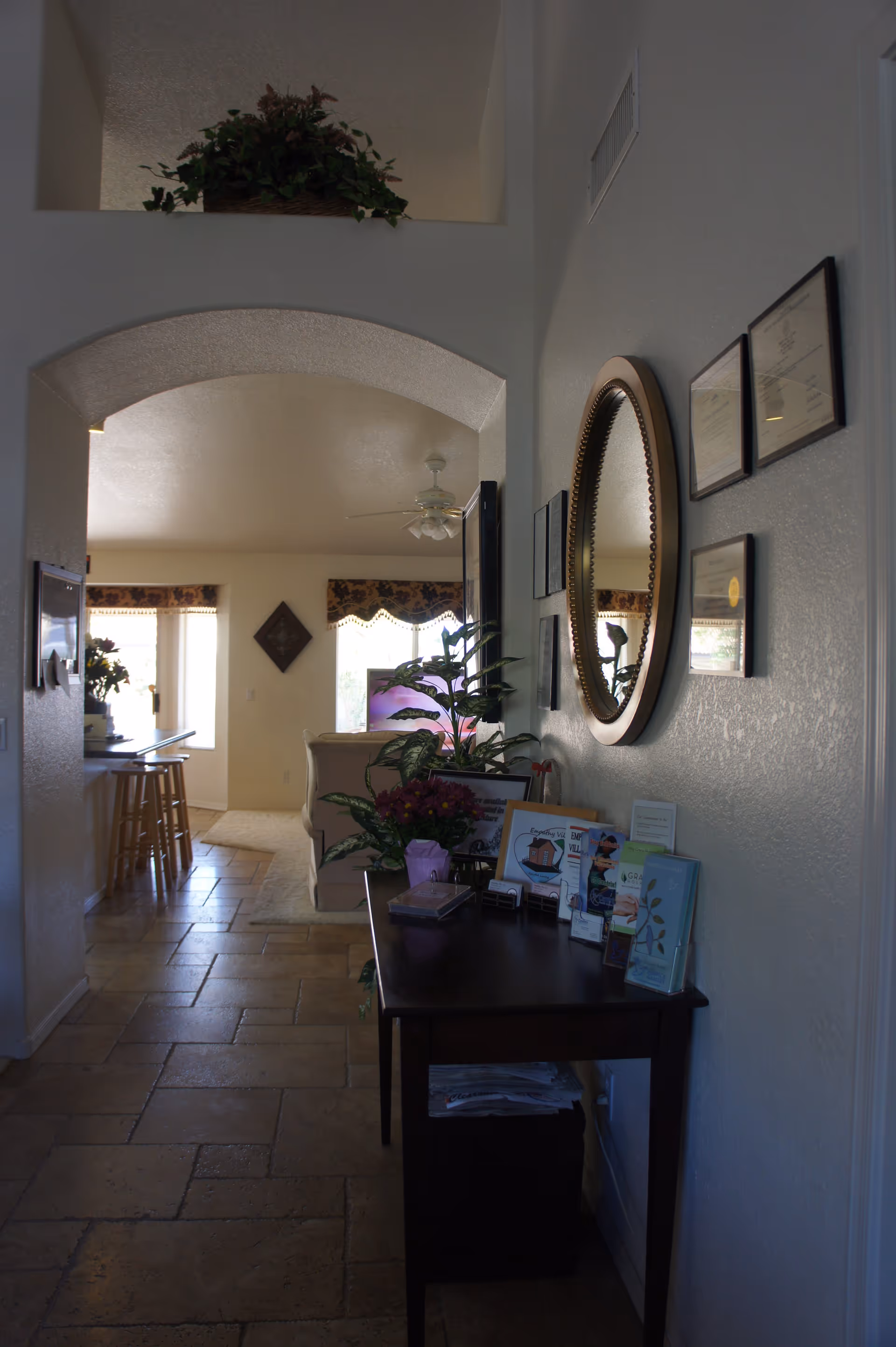 A tiled hallway leading into a living room with a console table topped with plants, brochures and a round mirror on the wall.