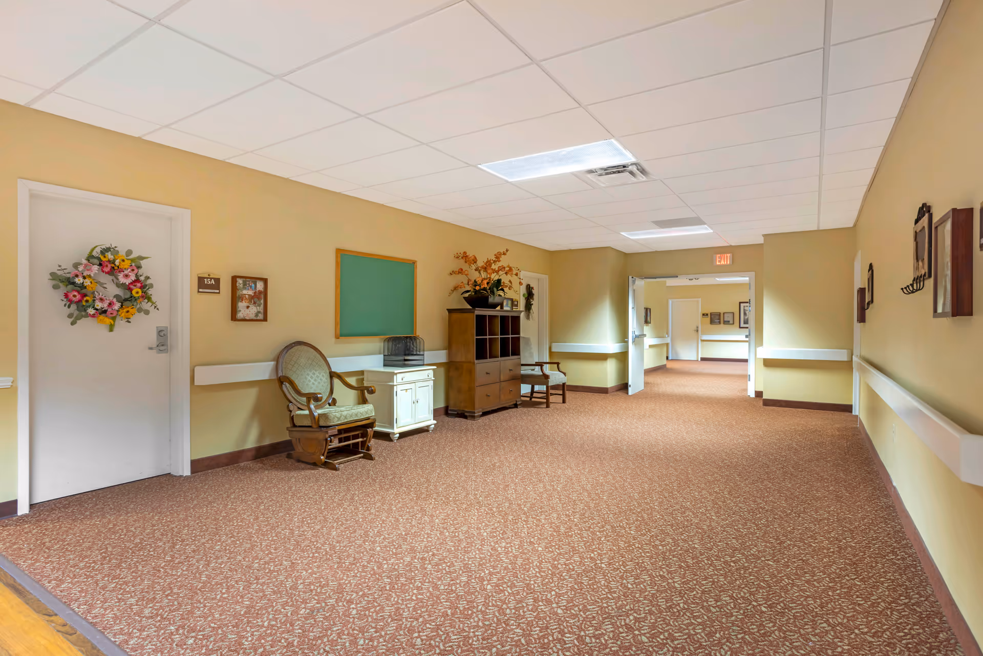 Carpeted interior hallway in a senior living facility with chairs, a cabinet, a decorated door, and an exit sign.